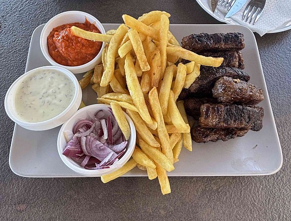 Porce čevapčiči s hranolkami, doplněná o misku s nakrájenou cibulí a dvěma miskami s omáčkami