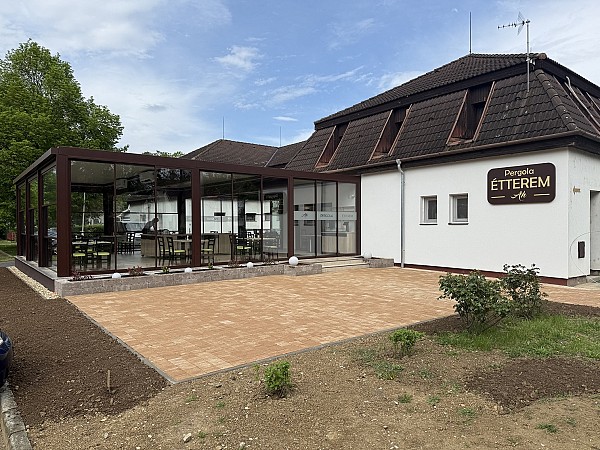 Moderní budova s bílou fasádou a hnědou střechou, u které je terasa se zastřešeným posezením