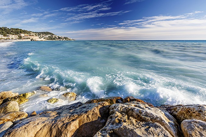 Mořské vlny narážející na skalnaté pobřeží pod modrou oblohou s oblaky