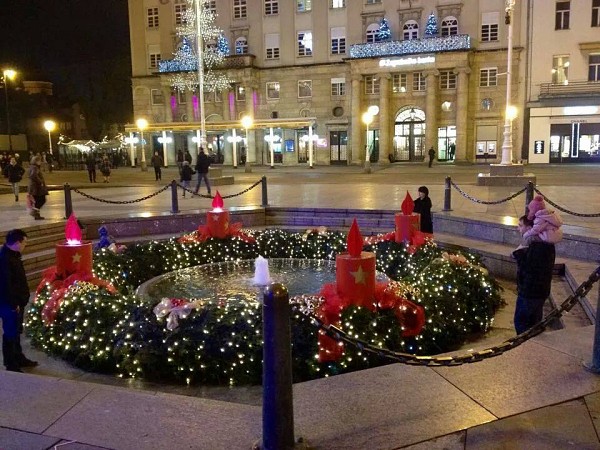 Adventní věnec se zapálenými svíčkami na náměstí v Záhřebu, obklopený lidmi při večerním osvětlení