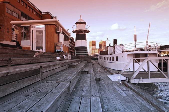 Přístavní molo v Oslu s dřevěnou promenádou, menším majákem a kotvícími loděmi za večerního světla