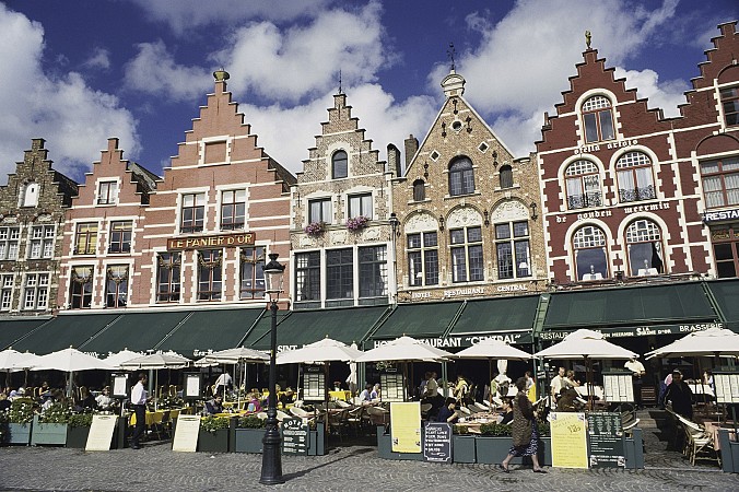 Historické barevné domy s tvarovanými fasádami na náměstí Markt v Bruggách, před nimi venkovní kavárny se slunečníky