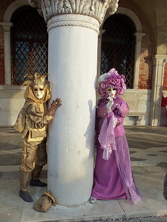 Dvě postavy v karnevalových kostýmech, zlaté a růžové barvy, stojí u kamenného sloupu před historickou budovou