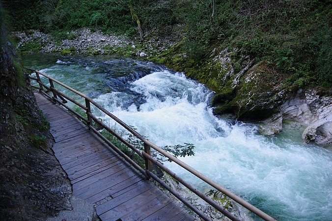 Dřevěná lávka se zábradlím podél řeky Radovna v soutěsce Vintgar ve Slovinsku, voda hučí přes balvany a vytváří bílé peřeje