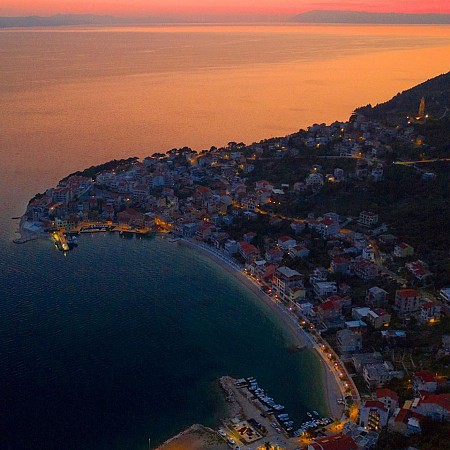 Pobřežní město Igrane s dlouhou pláží a zálivem, osvětlené oranžovým světlem zapadajícího slunce