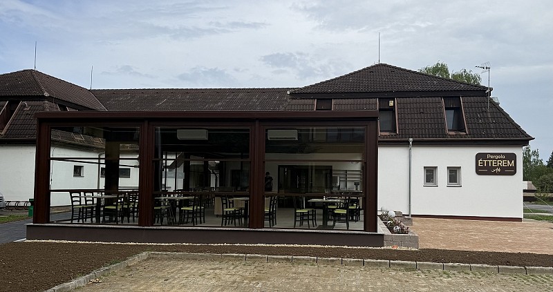 Budova s tmavou střechou a bílými zdmi má krytou pergolu se stoly a židlemi pro venkovní posezení