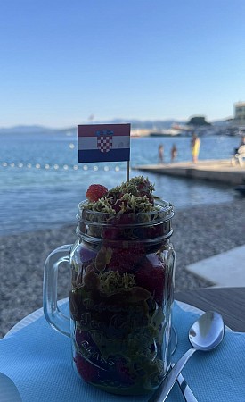 Ve sklenici jsou jahody obalené v čokoládě ve stylu Dubai Chocolate, podávané na stole u moře