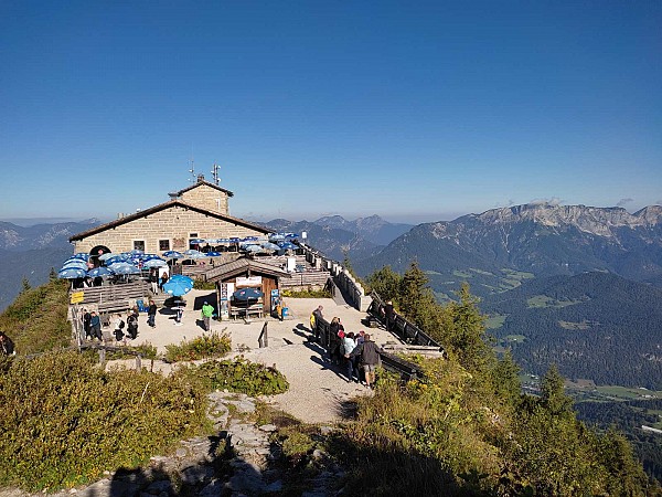 Horská budova s terasou a modrými slunečníky stojící na vrcholu s panoramatickým výhledem na okolní hory