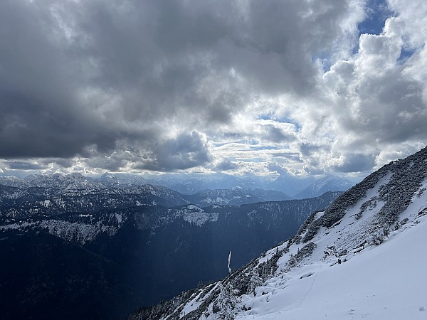 Sníh pokrývá horský svah, odkud je vidět hluboké údolí a vzdálené vrcholky hor pod zataženou oblohou