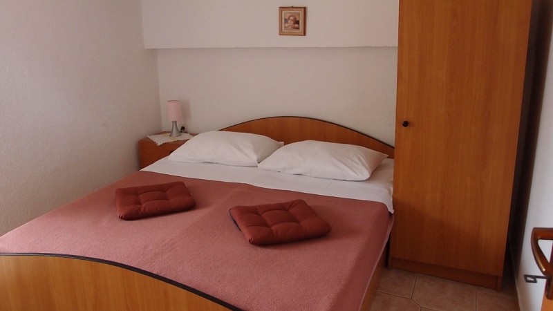 Pokoj s dřevěnou manželskou postelí povlečenou růžovým přehozem, dvěma polštáři a složenými ručníky