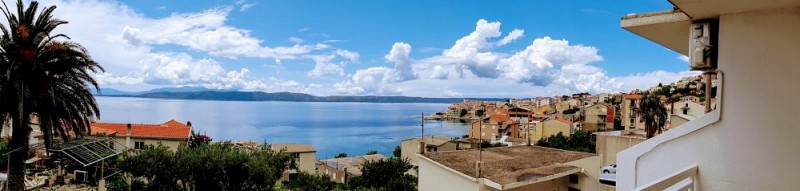 Výhled z balkonu na moře s červenými střechami domů a palmou