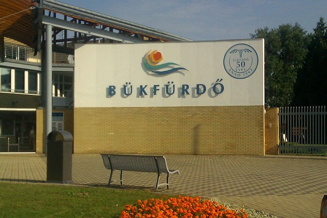 Vstupní cedule termálních lázní Bükfürdő s logem areálu a lavičkou poblíž
