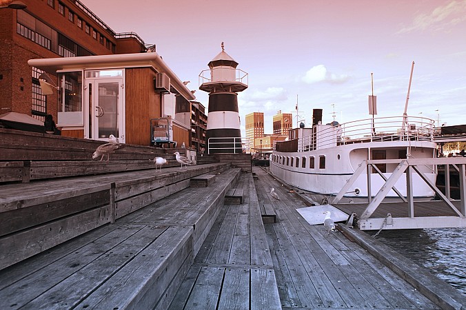 Přístavní molo v Oslu s dřevěnou promenádou, menším majákem a kotvícími loděmi za večerního světla