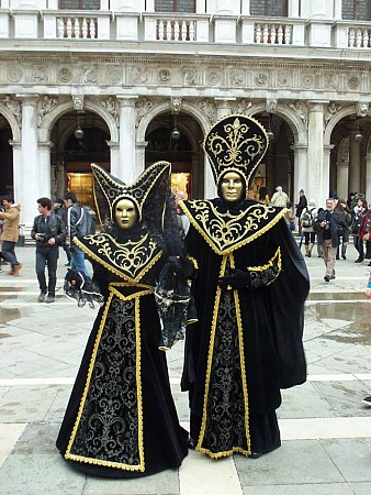 Dva lidé stojí v historických kostýmech s černozlatými maskami na benátském náměstí