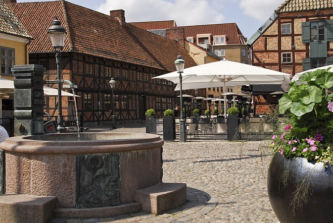 Malebné náměstí v Malmö se starobylými hrázděnými domy, venkovním posezením pod velkým slunečníkem a kašnou