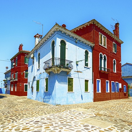 Ulice s barevnými domy – světle modrý dům vpředu a červené domy po stranách – pod jasně modrou oblohou