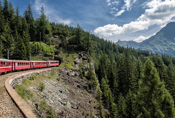 Červený vlak projíždí po horské trati mezi zelenými lesy a skalami