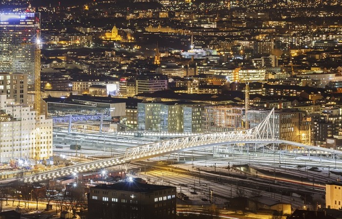 Moderní budova opery v Oslu s bílými šikmými plochami osvětlená v noci, svítící město s vysokými kancelářskými domy