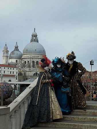 Tři lidé v barokních karnevalových kostýmech a maskách stojí na mostě
