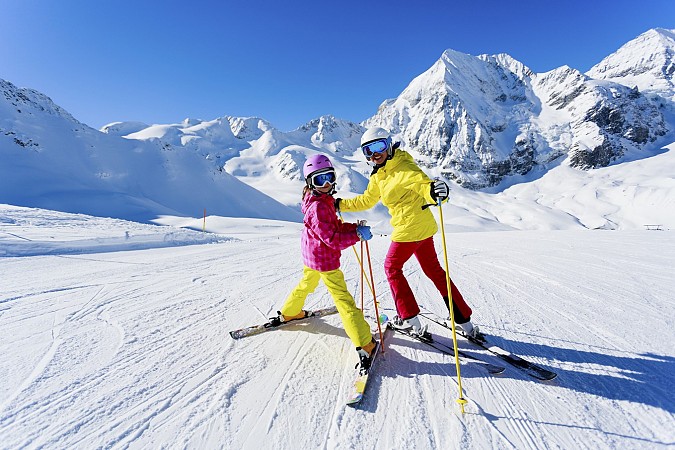 Dva lyžaři v barevném oblečení stojí na zasněžené sjezdovce obklopené vysokými horami