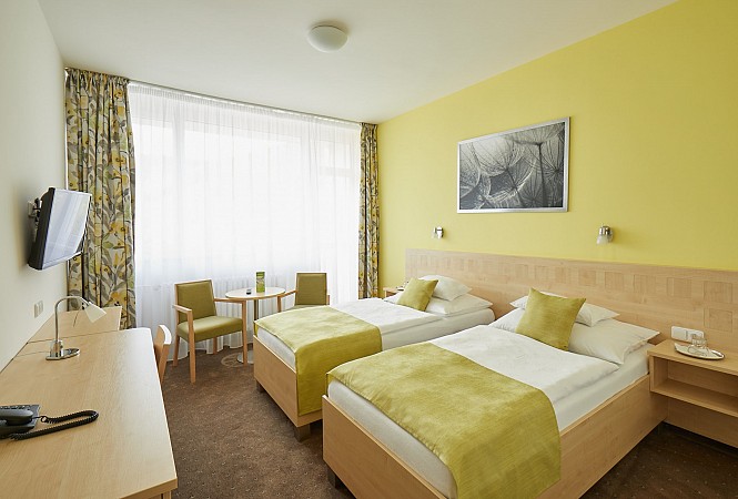 Světlý hotelový pokoj se dvěma postelemi, žlutými doplňky a posezením u okna