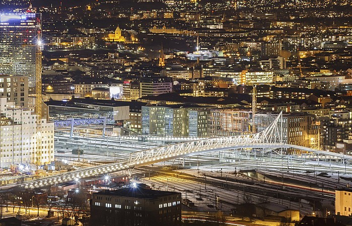 Moderní budova opery v Oslu s bílými šikmými plochami osvětlená v noci, svítící město s vysokými kancelářskými domy