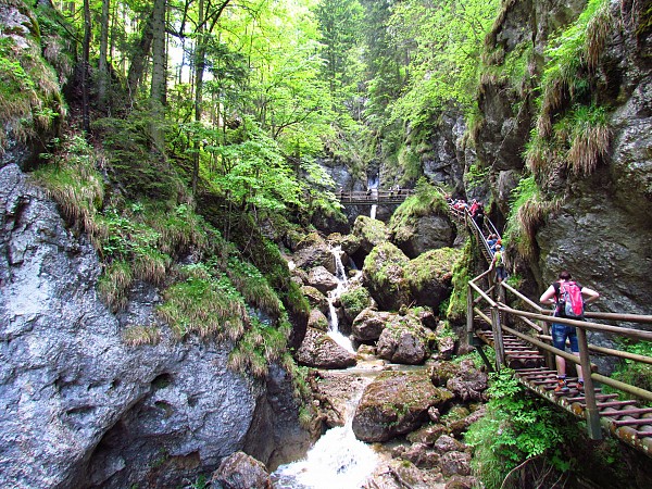 Úzká soutěska se skalami, malým potokem a dřevěným chodníkem, po kterém procházejí lidé