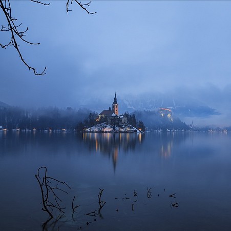 Ostrov na jezeře Bled ve Slovinsku s osvětleným kostelem Nanebevzetí Panny Marie obklopeným mlhou